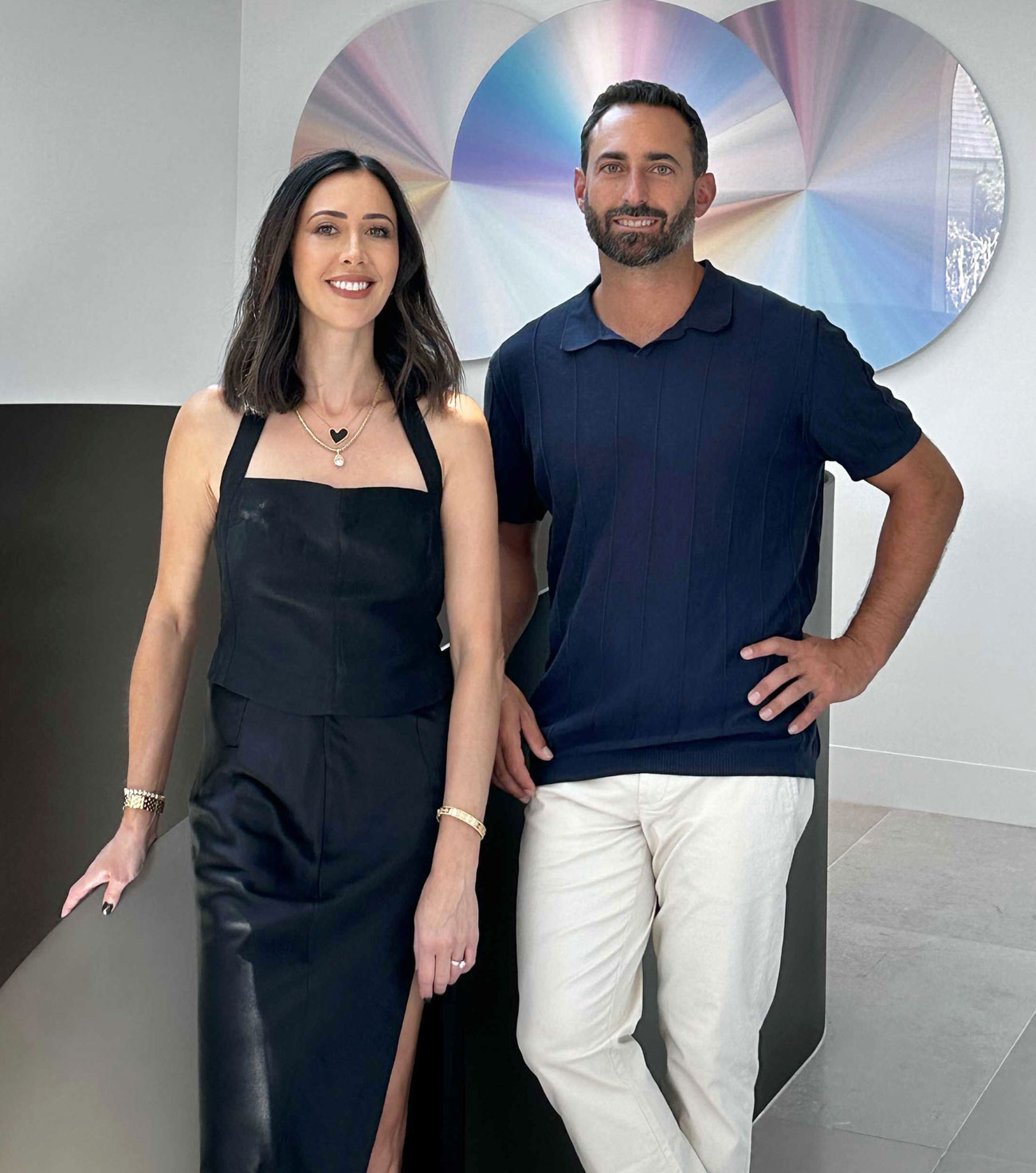 Nina Maya and Colin Sternberg stand side by side in front of an abstract, iridescent wall art piece. Nina, on the left, is wearing a sleeveless black dress with a subtle sweetheart neckline and bracelets on both wrists. Colin, on the right, sports a dark blue collared shirt with rolled-up sleeves and off-white pants. They are both smiling subtly and appear relaxed.